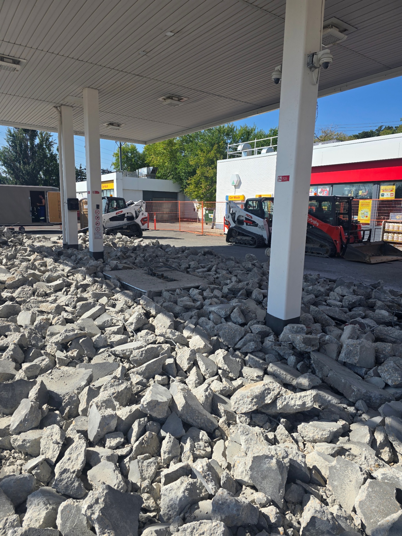 Concrete demolition under a gas station canopy with skid steers removing the old fuel island slab